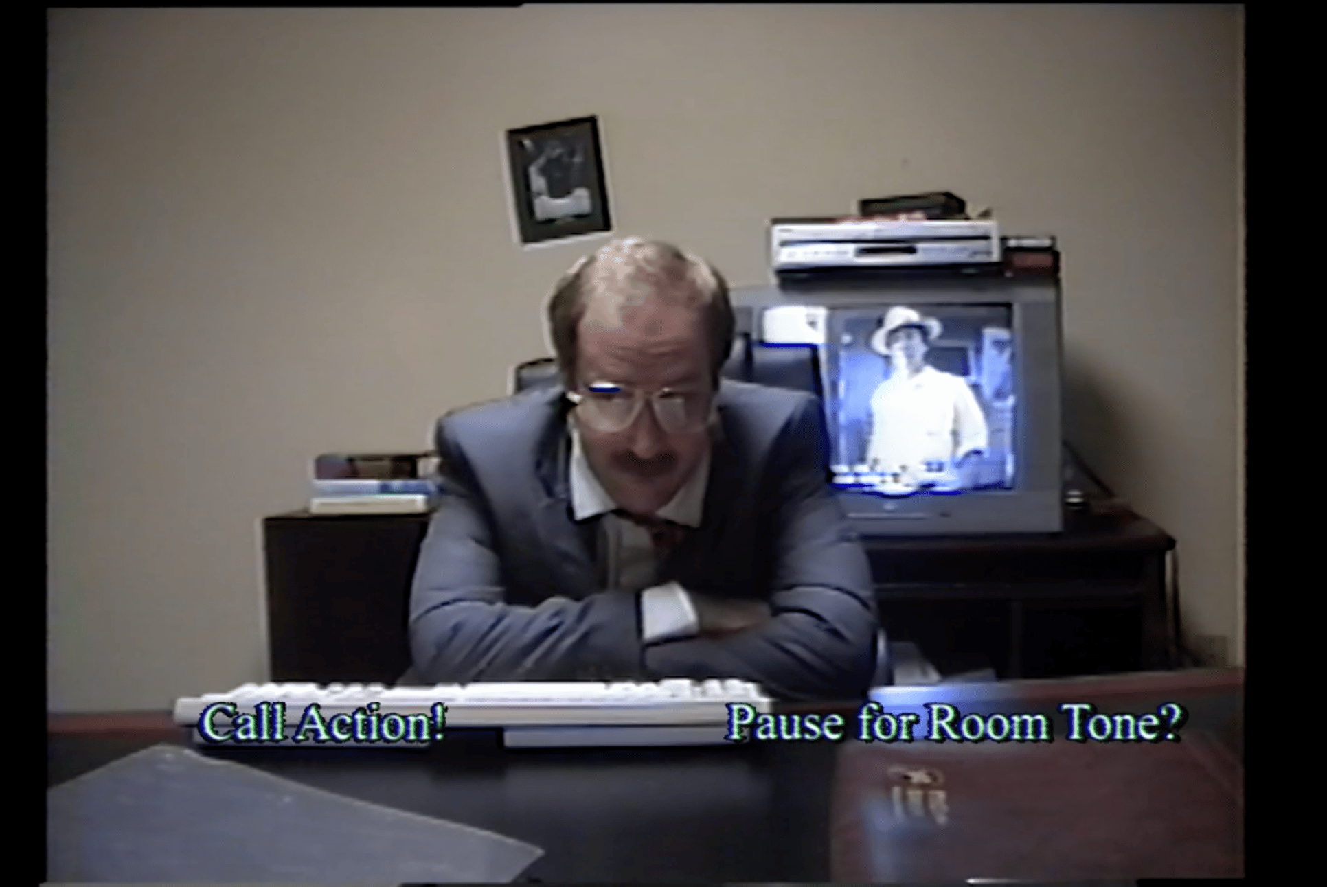 VHS camera footage - a man wearing glasses and a suit and sits at a desk. There is a TV behind him. Two choices are given on screen: "Call for action", "pause for room tone"