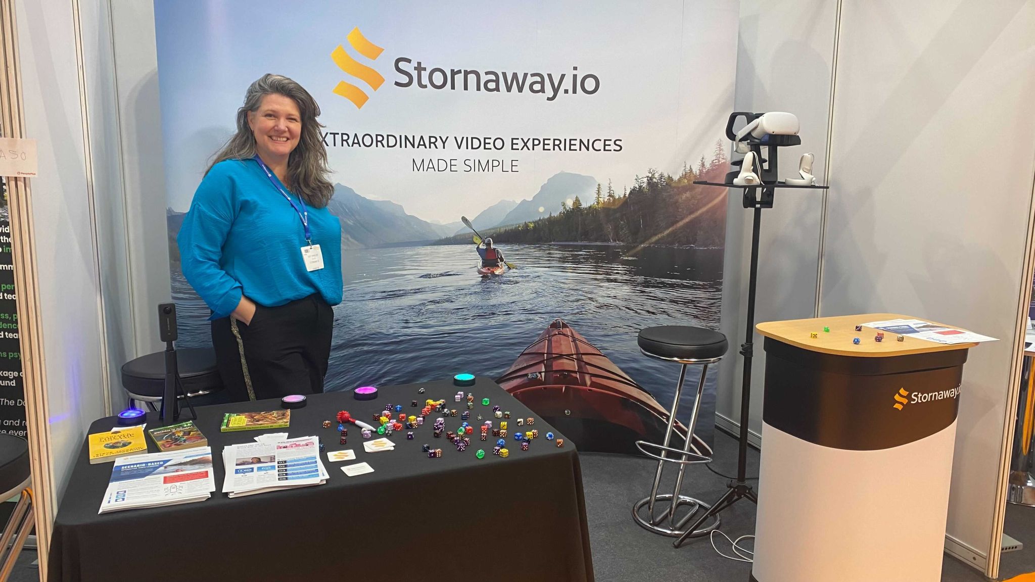 Kate Dimbleby - a lady with long brown hair wearing a blue shirt - stands in front of a poster for Stornaway.io at a conference
