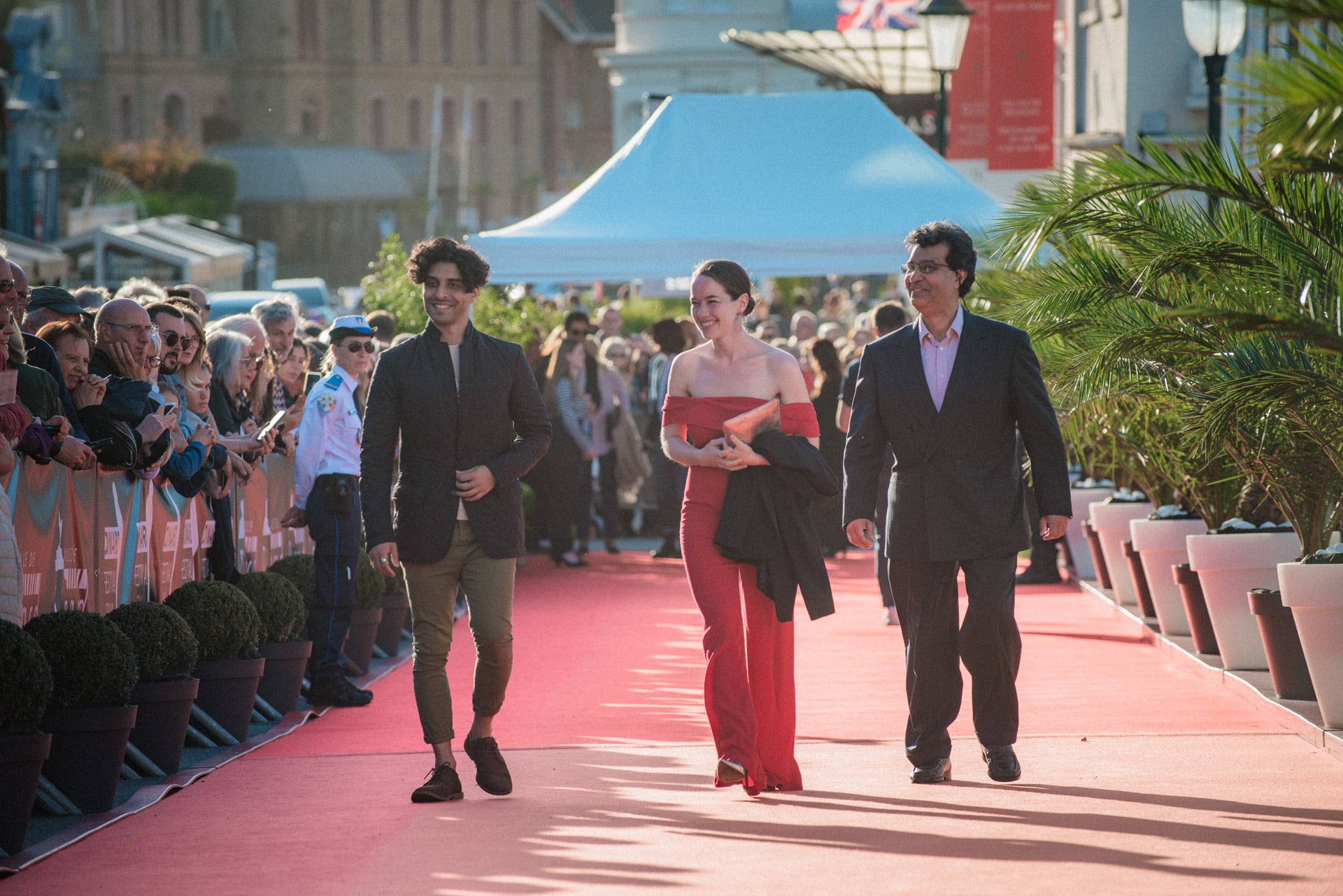 A red carpet scene. A man in a dark blazer and light trousers walks with a lady in a long red dress and another man in a dark suit. There are crowds and photographers.