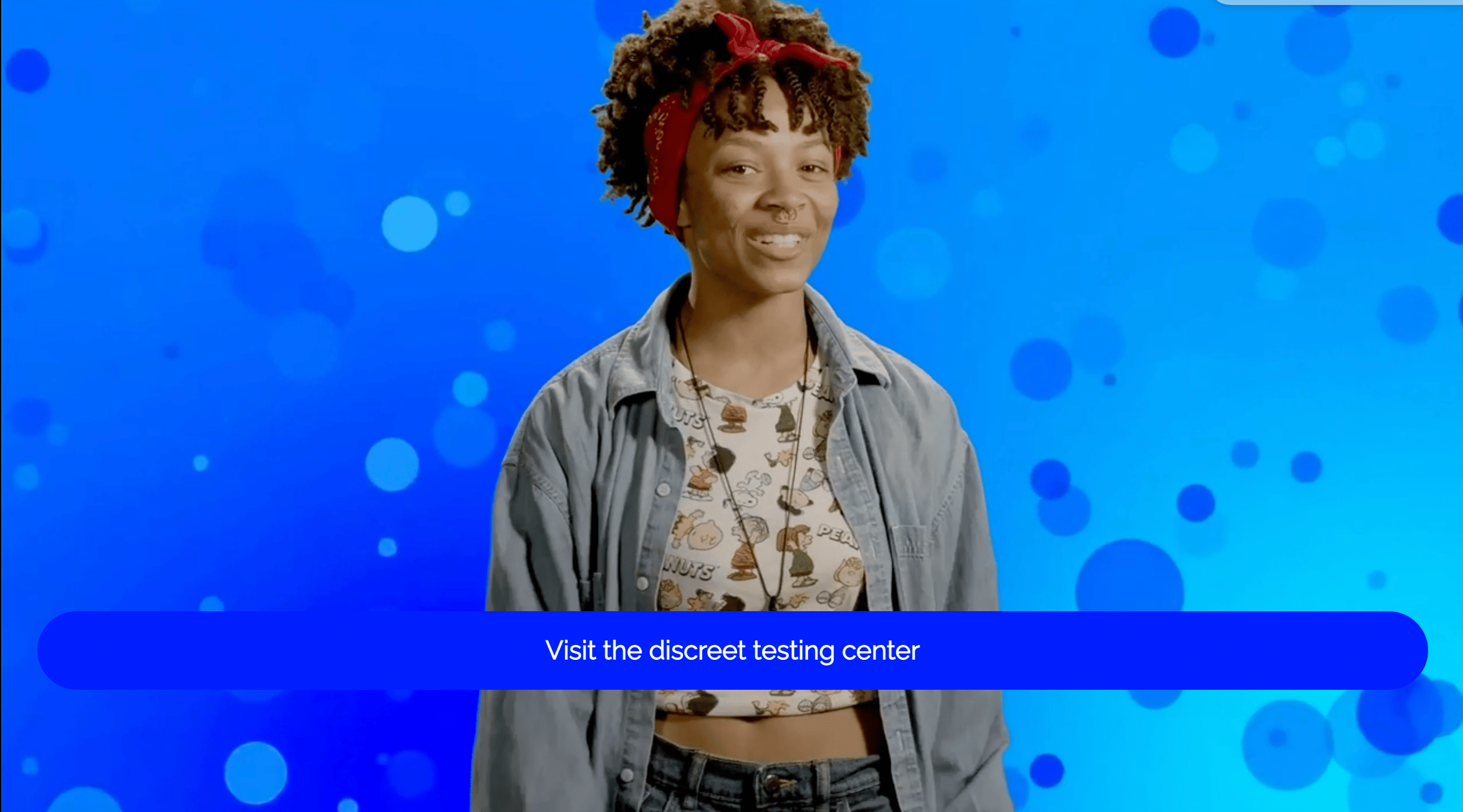 DatingForward - behaviour changing interactive video - A woman stands in front of a blue background. She is wearing a red scarf in her hair, a long open shirt and a crop t shirt. She is talking to camera. A blue box with white writing contains the words: "Visit the discreet testing centre"