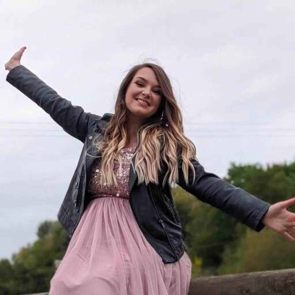 Gemma Twomey - Head of Product at Stornaway.io. A woman wearing a pink floaty dress and a black leather jacket holds her arm out. She is smiling and look at the camera.