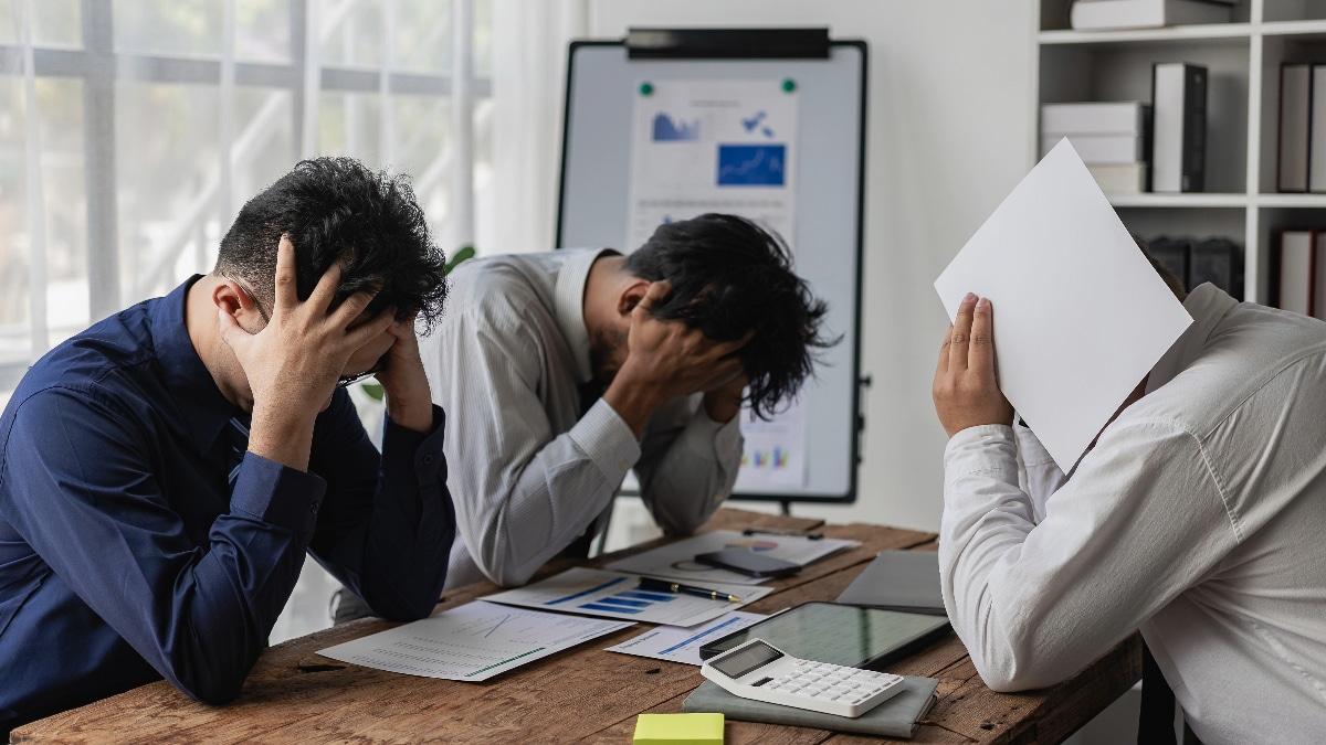 persuade your boss to buy new software - Three men sit with their heads in their hands in an office. two are wearing white shirts. One is wearing a navy shirt. One of the men in the white shirts sits opposite the other two, he is holding a sheet of paper to cover his face.