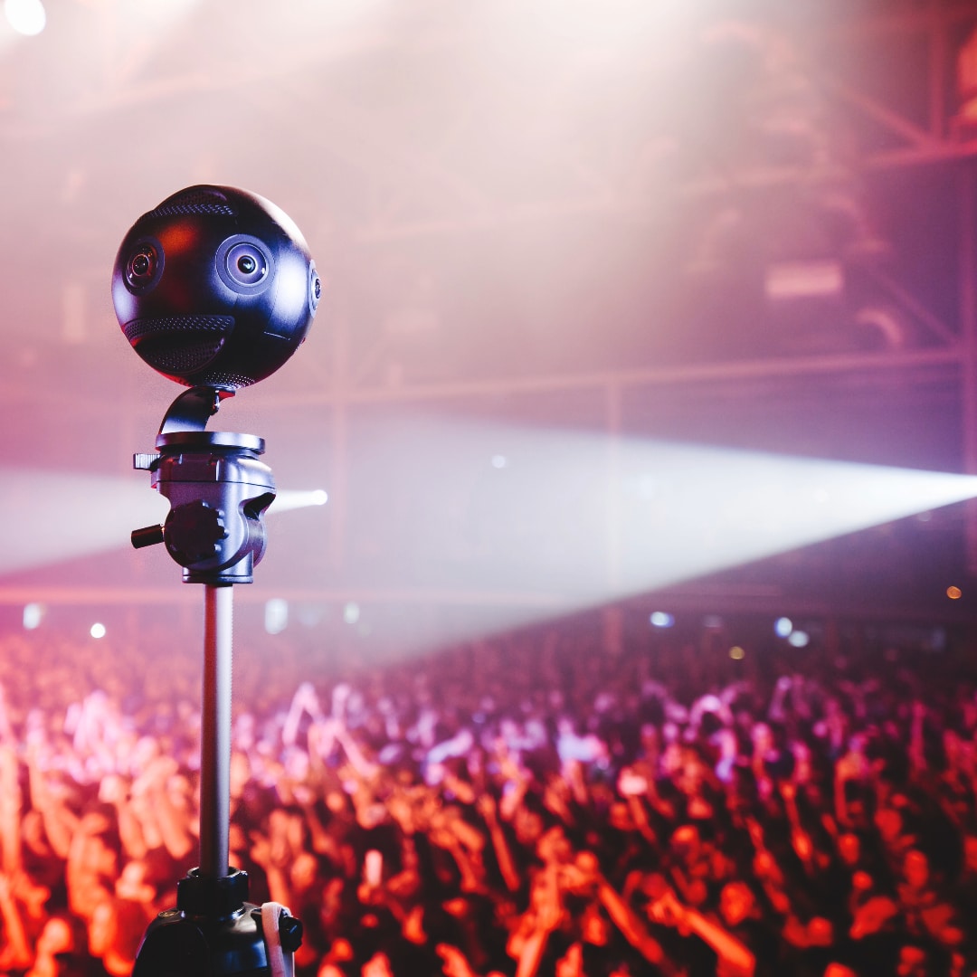 360 camera at a gig - a 360 camera is placed above the crowd at a gig. The people are blurred in the background with raised arms. Red, orange and purple lights shine.