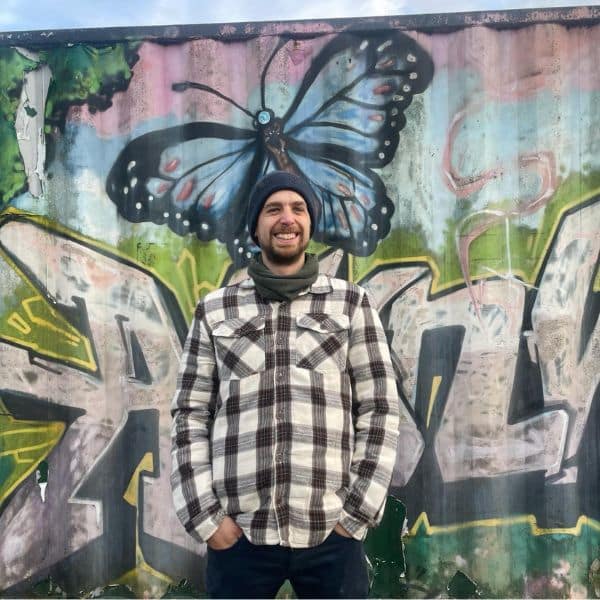 Brock Horning - Senior Copywriter - A white, bearded, smiling man stands in front of a graffitied metal container. He wears a blue beanie and chequered shirt.