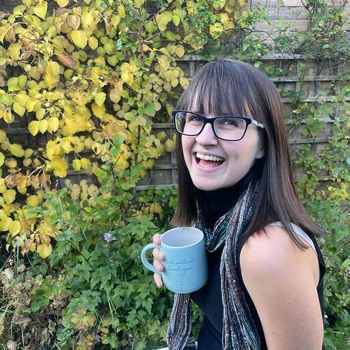 Abbie Horning - head of communications - a woman with darrk hair and a fringe smiling in a garden with a blue mug and a colourful scarf