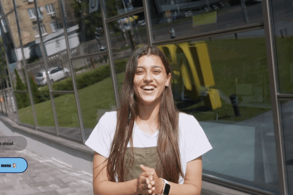 Interactive Tour - created using Stornaway's Design Editor - A young woman with long brown hair smiles and clasps her hands together while standing outside a modern glass building on a sunny day. She is wearing a white T-shirt under green overalls and a smartwatch. Two interactive buttons labeled “>> Skip ahead” and “Back to menu 🏠” appear on the left side of the image, indicating it was made using Stornaway’s interactive video design editor.