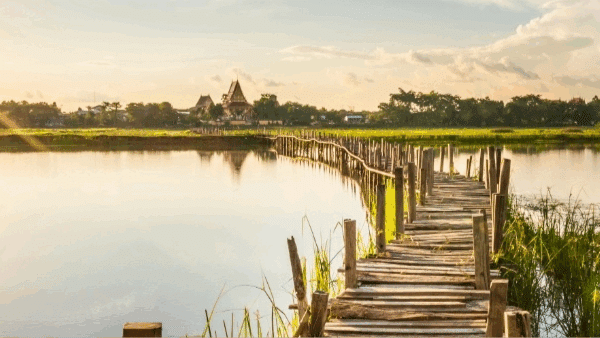 Event title: “The Business of Interactive Video series: Interactive Learning — How 4 learning experts built businesses with interactive video.” A long, rustic wooden footbridge stretches across a calm lake toward a distant cluster of buildings and trees, glowing under a warm sunset sky. Lush grasses and reflections shimmer in the water, creating a peaceful, inviting atmosphere.