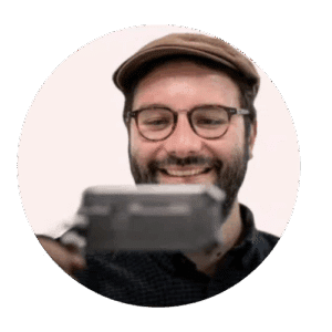 Dr Stefano Odorico – Stornaway Academic Summit: Exploring Polyphonic Documentary with Stornaway. A man with a beard and glasses is smiling while holding a camera or recording device. He’s wearing a brown flat cap and a dark checkered shirt, with a plain light background behind him.