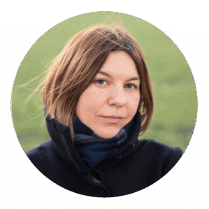 Lena Dobrowolska – Stornaway Academic Summit: Exploring Polyphonic Documentary with Stornaway. A woman with light brown hair and blue eyes is standing outdoors, wearing a dark coat and a navy scarf. She has a calm, thoughtful expression, with a soft-focus green background of grass behind her.