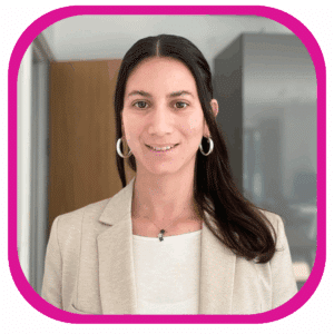 Micaela Valdizan Woodman - Professional headshot of a woman with long dark hair, wearing a light beige blazer over a white top, silver hoop earrings, and a delicate necklace. She is smiling slightly and standing indoors in a modern office setting with neutral-toned walls and cabinetry softly blurred in the background.